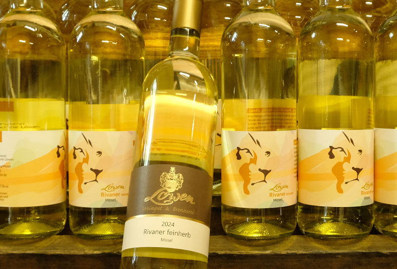 Reihe von Flaschen Rivaner feinherb nebeneinander. Die Etiketten sind in unterschiedlichen Postionswinkeln zu sehen. In der Mitte ist eine schrägplatzierte Flasche Rivaner mit dem bisherigen Etikett.