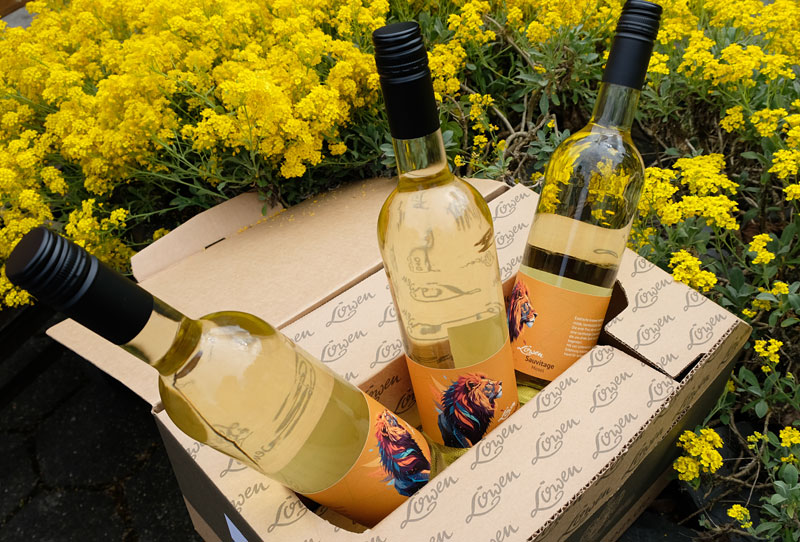 Drei Weinflaschen in Karton, der im Grünen platziert ist. Gelbe Blumen umgeben den Karton.